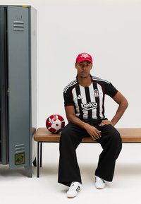 Mustvalge triibulise spordisärgi punaste detailidega, mustad laiad püksid, valged kingad, punane müts. Läheduses punane ja valge jalgpall, taga hall lukustuskap.