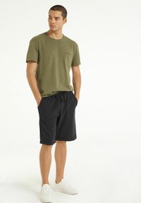 Olivově zelené bavlněné tričko s krátkým rukávem a kapsou na hrudi, v kombinaci s černými šortkami na šňůrku a bílými teniskami.