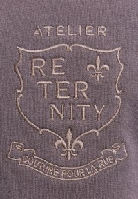 Broderie sur tissu représentant un blason avec l'inscription "RETERNITY", des symboles de fleur de lys, "ATELIER" au-dessus, et "COUTURE POUR LA RUE" sur un ruban en dessous.