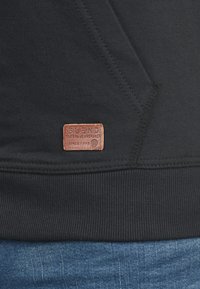 Sudadera negra con ribete en el dobladillo, costuras laterales y una etiqueta de cuero marrón con el texto "BLEND" en relieve, que forma parte del diseño.