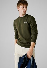 Olijfgroen sweatshirt met ribbelboorden en halslijn, voorzien van het witte "Red Bull Racing" logo. Model houdt een spijkerjack in één hand.