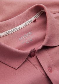 Rosa bomullspiké med krage, texturerad tyg, tre knappar och en etikett synlig i halsringningen som anger "100% ren bomull."