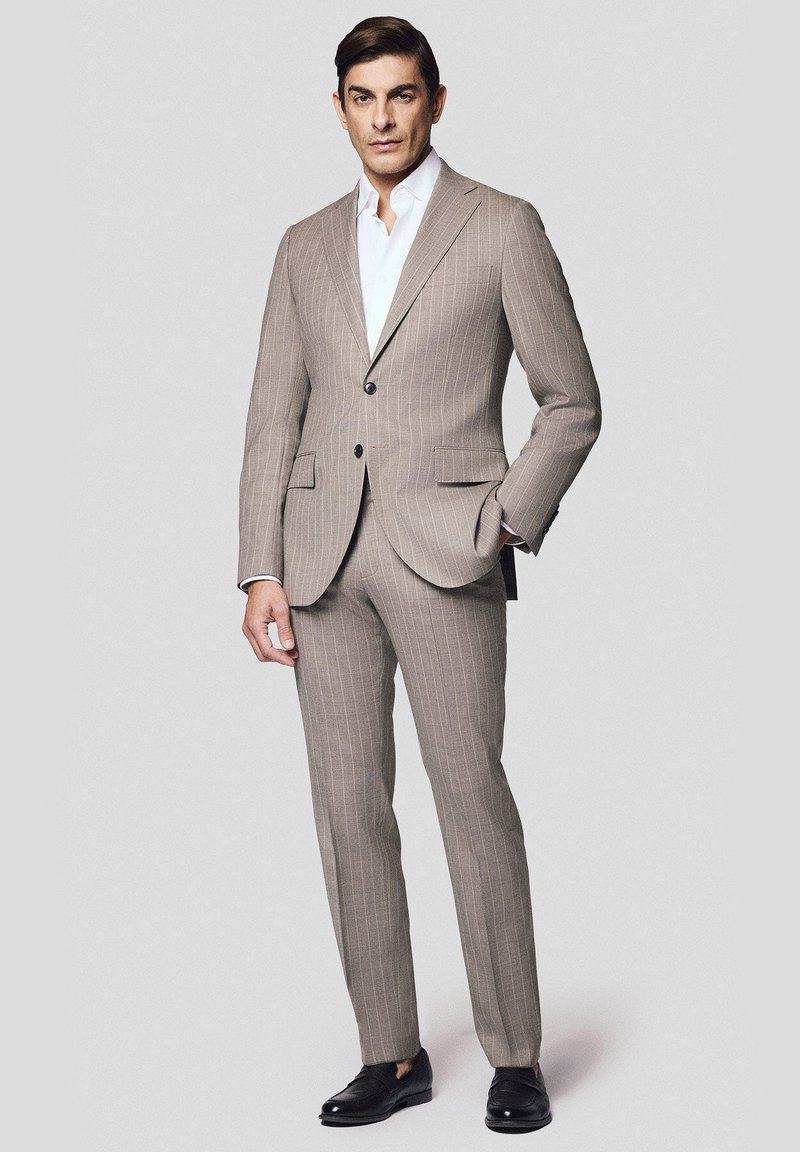 Brązowy garnitur w prążki wykonany z teksturowanej tkaniny, z dwoma przednimi kieszeniami, klapami w kształcie litery V i zapięciem na jeden guzik. Połączony z białą koszulą i czarnymi butami.