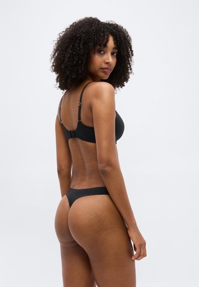 Femme aux cheveux bouclés portant un soutien-gorge noir et une culotte string, regardant par-dessus son épaule sur un fond clair uni.