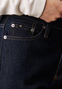 Granatowe dżinsy z kontrastowym ś stitchingiem, metką CK oraz metalowymi akcentami w talii i kieszeniach. Widoczna teksturowana tkanina.