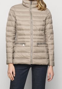 Beige vadderad jacka med hög krage, framdragkedja och två fickor med dragkedja. Horisontellt quiltat tyg ger textur åt designen.