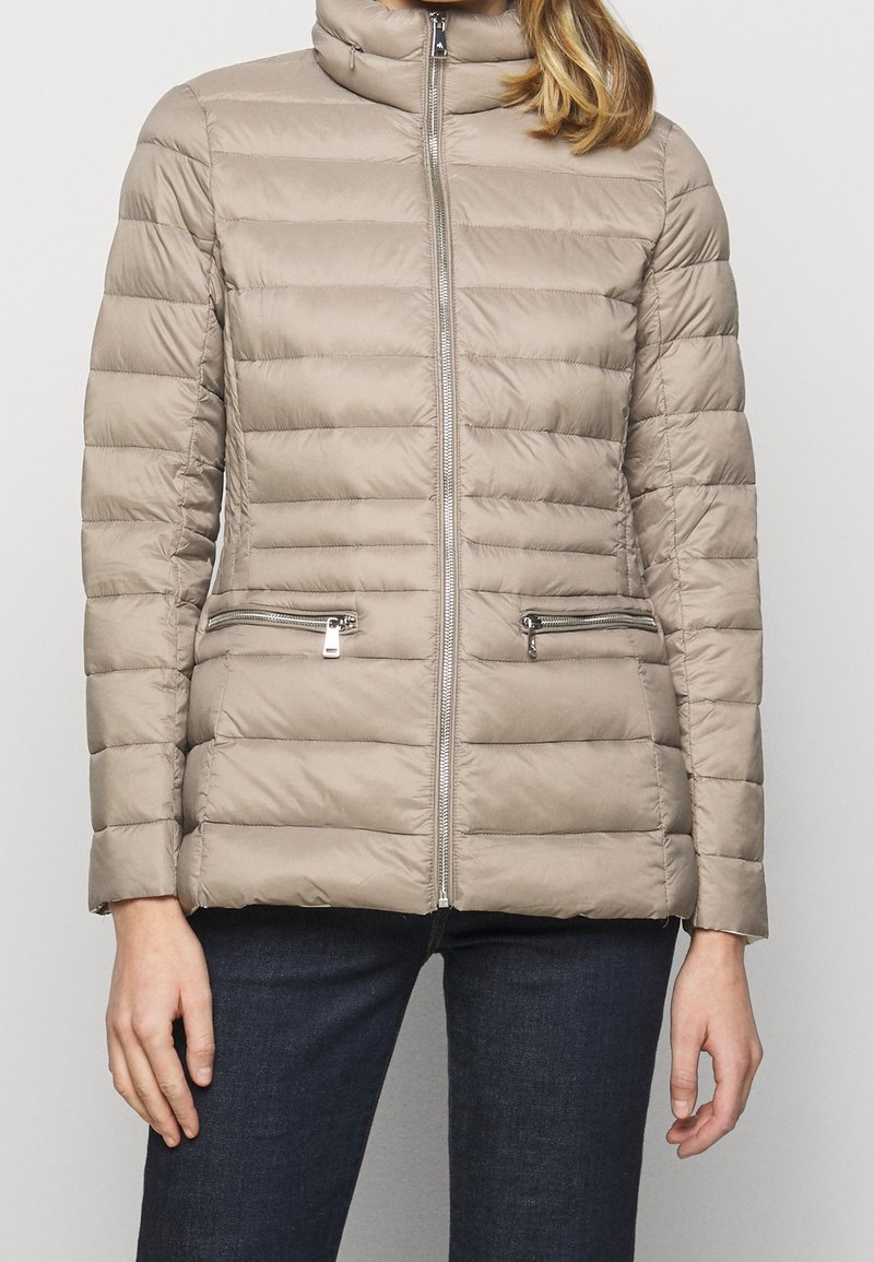 Beige vadderad jacka med hög krage, framdragkedja och två fickor med dragkedja. Horisontellt quiltat tyg ger textur åt designen.