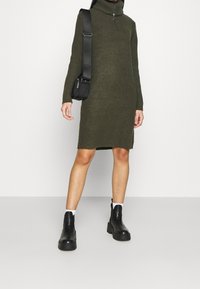 Strikket grøn turtleneck kjole med ribbet tekstur, der når midt på låret. Båret med sorte platform ankelsko og en lille sort crossbody taske.