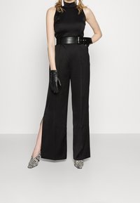 Top sin mangas negro con cuello alto, pantalones anchos negros con aberturas laterales, guantes negros y zapatos de punta a rayas blanco y negro.