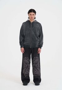 Sudadera gris con cremallera y acabado texturizado, adornada con remaches de metal. Combinada con pantalones vaqueros negros de pierna ancha que presentan detallados patrones de serpiente.