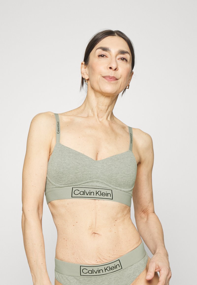 Gaiši pelēks Calvin Klein bralete, kas izgatavota no mīksta auduma, ar regulējamiem siksnām un ar logo joslu. Gluda tekstūra un bezšuvju dizains.