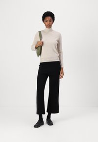Bež pulover s turtleneck ovratnikom, črne široke hlače in črne loafers čevlje. Drži zeleni nahrbtnik, stoji ob navadni beli ozadju.