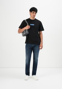 Jonge man in een zwart Tommy Jeans T-shirt, blauwe spijkerbroek, witte sportschoenen, met een grijze schoudertas over één schouder tegen een witte achtergrond.