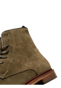 Stivale in camoscio verde oliva, con punta rotonda, lacci in pelle marrone e tacco in legno impilato con accenti marroni.