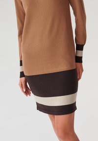 Abito lavorato a maglia con strisce marroni, crema e nere. Maniche lunghe con polsini a coste. Texture liscia, design aderente e taglio al ginocchio.