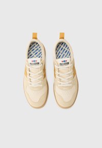 Cream sneakers with tan suede accents, white laces, and a logo patch. Interior features a blue patterned lining. Flat rubber sole.