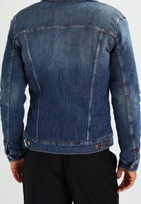 Blå denimjacka med smal passform, knappade ärmslut och kontrasterande sömmar. Baksidan har en yoke och subtil blekning vid axlarna.