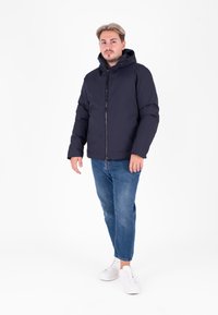 Giacca imbottita blu marina con cappuccio, zip frontale e polsini elastici. Indossata con jeans azzurri e sneakers bianche. Design semplice e funzionale.