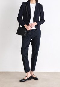 Mörkblå Blazer, vit t-shirt med logotyp, skräddarsydda mörkblå byxor och svarta ballerinaskor. Liten svart axelremsväska. Släta texturer.