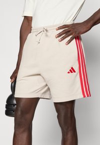 Μπεζ αθλητικό σορτς με κόκκινες ρίγες στα πλαϊνά, ελαστική ζώνη μέσης με κορδόνι και μικρό κόκκινο λογότυπο στο αριστερό πόδι.