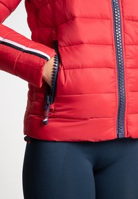 Rød dunjakke med lynlåslukning foran og sidelommer. Har marineblå detaljer og striber ved manchetterne samt et struktureret, polstret udseende.