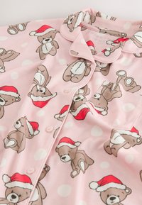 Pink pajama top with cream polka dots and a teddy bear print wearing red hats. Features a collar and button closure. Soft fabric texture.