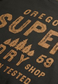 Camiseta negra de tela con letras doradas desgastadas. El texto incluye "OREGON SUPER DRY 59" y un gráfico de un árbol sobre la palabra "DRY."