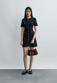 Γυναίκα με κοντό μαύρο φόρεμα με κουμπιά και τσέπες, κρατώντας μια σκούρα κόκκινη τσάντα, φορεί γκρι κάλτσες και μαύρα μοκασίνια, στέκεται μπροστά σε ένα απλό τοίχο.