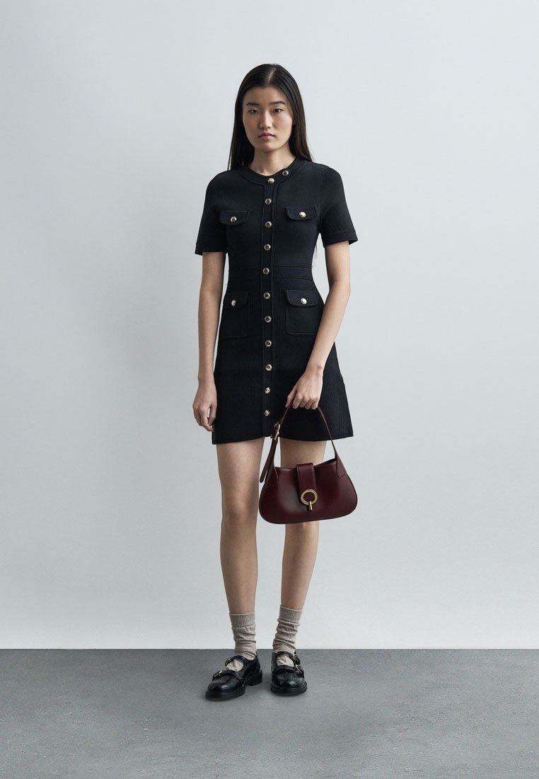 Mujer con vestido corto negro abotonado con bolsillos, sosteniendo un bolso rojo oscuro, calcetas grises y mocasines negros, de pie contra una pared lisa.