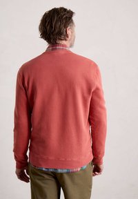 Felpa rossa con scollatura rotonda, polsini e orlo a costine. Indossata sopra una camicia a quadri multicolore. Tessuto misto cotone, vestibilità comoda.
