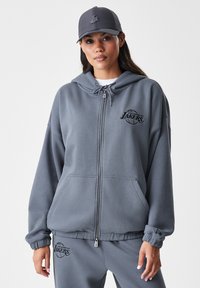 Felpa grigia con zip, cappuccio regolabile, polsini a costine e tasche frontali; presenta il logo ricamato "Los Angeles Lakers" sul petto.