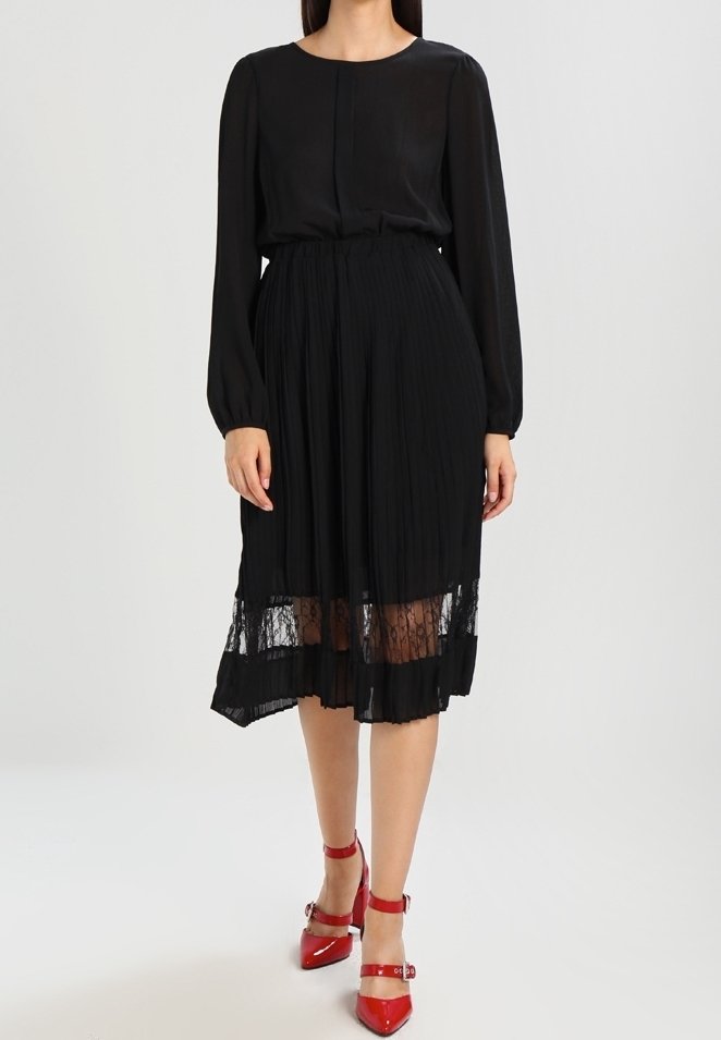 Femme portant une robe noire à manches longues avec un ourlet en dentelle transparente et des talons hauts rouges à bride à la cheville, debout devant un fond uni.
