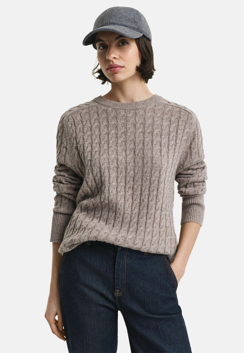 Kabelgebreide trui in zacht beige, met een ronde halslijn en geribde manchetten. Gecombineerd met donkerdenim jeans. Een grijze vilten pet completeert de look.