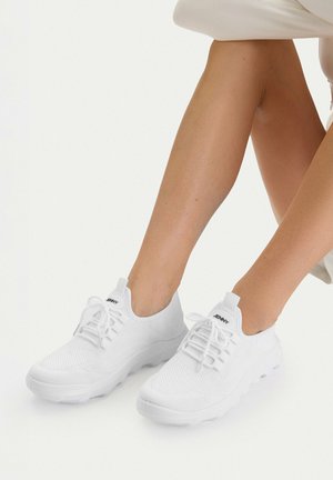 Weiße gestrickte Turnschuhe mit Schnürsenkeln, getragen an nackten Füßen, sitzend mit übereinandergeschlagenen Beinen vor einem schlichten weißen Hintergrund.