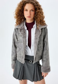 Graue Fake-Shearling-Jacke mit Kapuze und Taschen, die eine weiche Textur aufweist, kombiniert mit einem grauen Faltenrock und einem weißen Hemd mit Knöpfen.