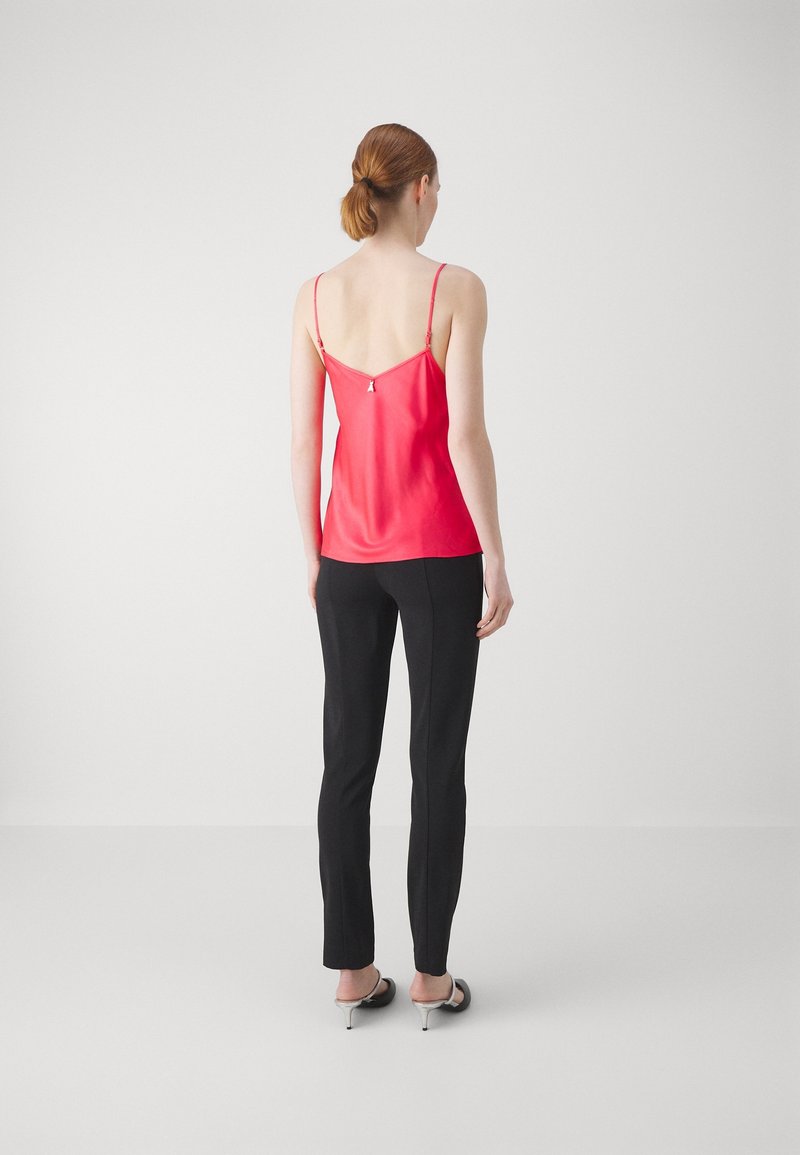 Mujer con cabello rojo recogido en una coleta baja, vestida con un top rojo sin mangas, pantalones negros ajustados y sandalias blancas con tacón, de pie frente a un fondo blanco liso.