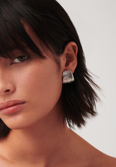 Γυναίκα με κοντά σκούρα μαλλιά που φοράει ασημένιο σκουλαρίκι σε σχήμα ανεμιστηράκι με υφή στο αριστερό αυτί της, σε απλό ανοιχτό φόντο.