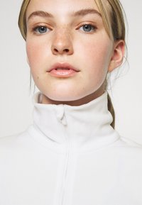 Weißes Fleece-Hochkragenoberteil mit einem Frontreißverschluss; glatte Textur und minimalistisches Design. Nahaufnahme des Halsbereichs und des Kragens.
