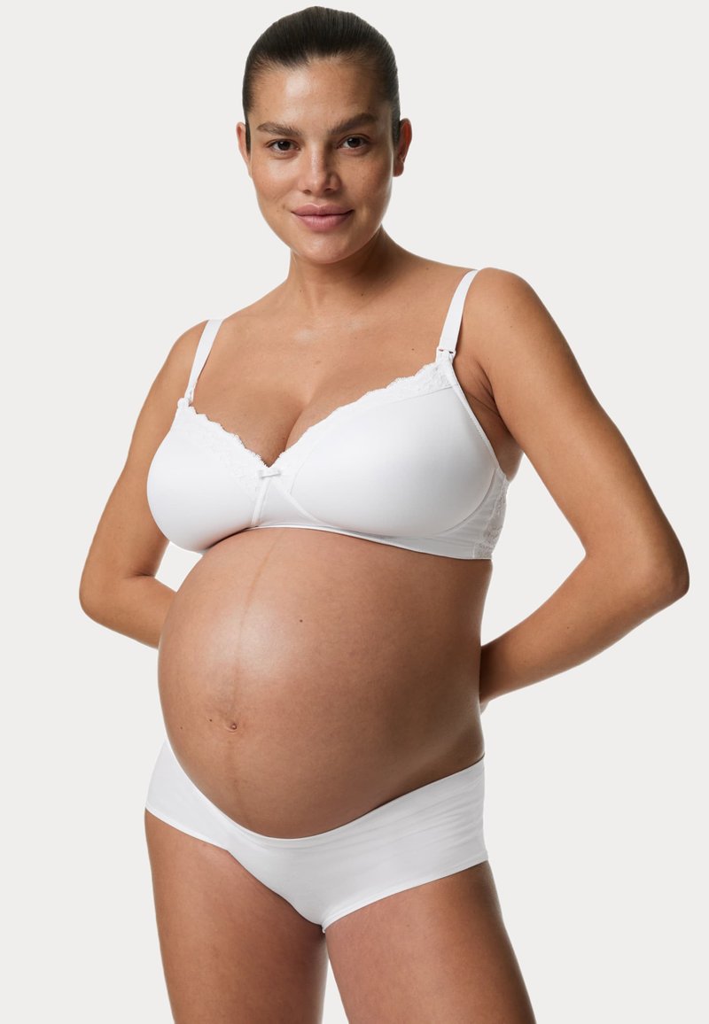 Hvid mødre-bh med blondertrim og justerbare stropper, parret med hvide trusser, båret af en gravid person. Glat stofstruktur.