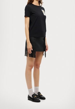 Frau, die ein schwarzes T-Shirt mit Brustpatch, einen schwarzen Fransenrock, weiße Socken und schwarze Loafers trägt, steht vor einem neutralen Hintergrund.
