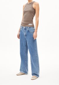 Kvinna iklädd en brun ärmlös topp, lösa ljusblå jeans och beige ballerinaskor som står mot en vit bakgrund.