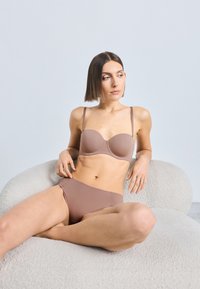 Svetlohnedá súprava spodnej bielizne s tvarovanou podprsenkou s nastaviteľnými ramienkami a vysokými nohavičkami, zobrazená na textúrovanej krémovej stoličke.