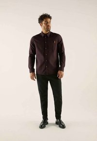 Man standing wearing dark purple button-up shirt, black pants, and black leather shoes on plain light background.