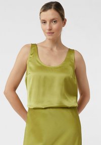 Vrouw met lichte huid en opgestoken haar, gekleed in een mouwloze top van limegroen satijn en een bijpassende rok tegen een effen achtergrond.