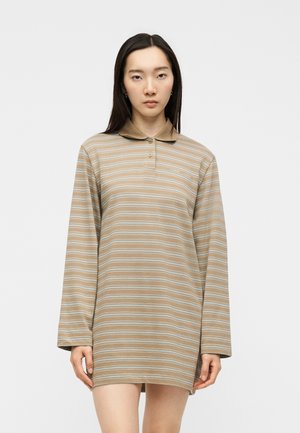 Kvinde med langt mørkt hår iført en beige og lyseblå stribet langærmet polokjole, stående mod en ensfarvet hvid baggrund.