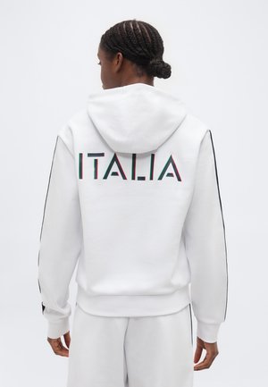 Person mit geflochtenen Haaren, die einen weißen Hoodie und eine weiße Hose trägt, mit der Aufschrift "ITALIA" in großen Buchstaben auf der Rückseite des Hoodies.