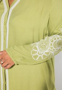 Camicia button-up verde chiaro con ricami floreali bianchi sulle maniche e sui bordi, caratterizzata da una texture morbida e una vestibilità rilassata.