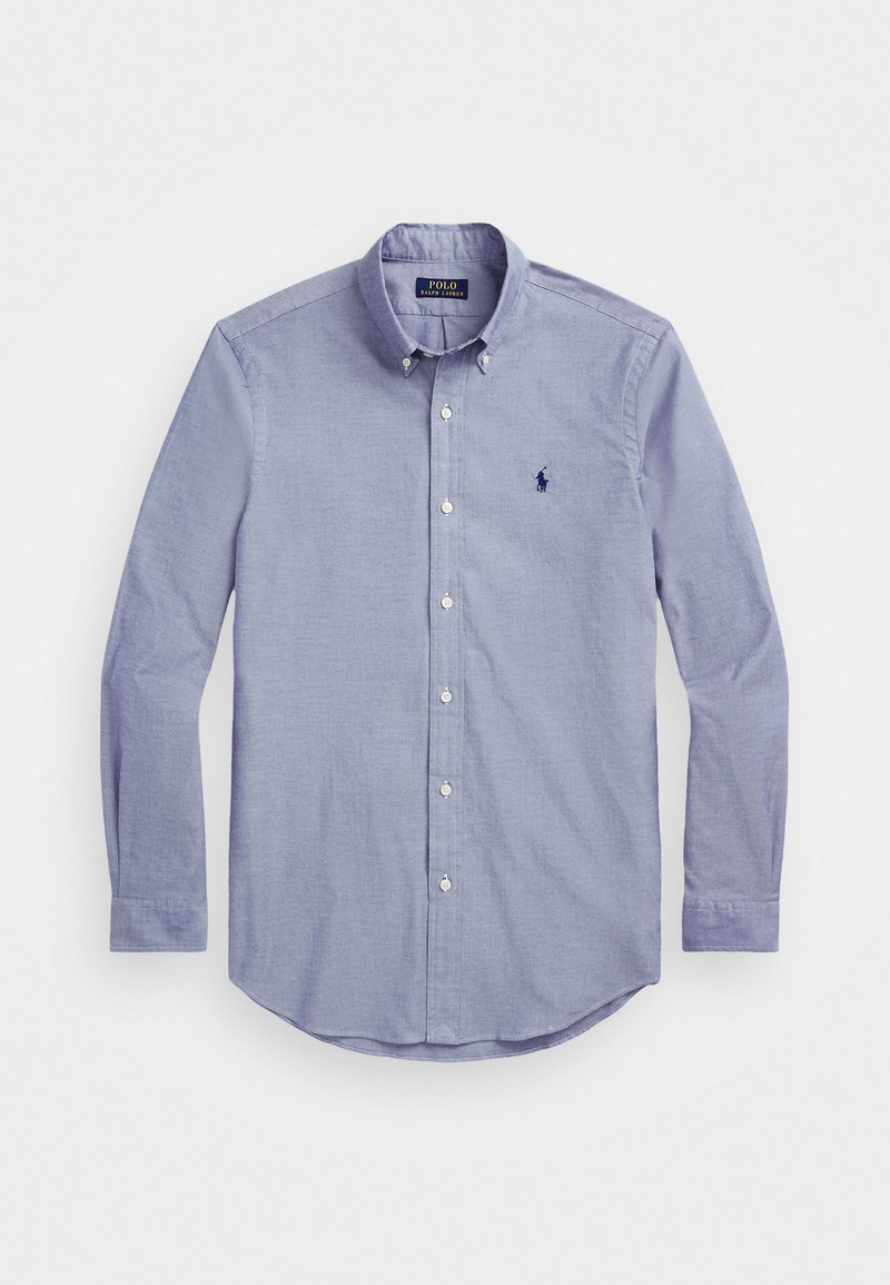 Šviesiai mėlynas marškinėliai su apykakle, ilgomis rankovėmis ir apvaliu apačiu; turi mažą išsiuvinėtą logotipą ant krūtinės. Pagaminta iš medvilnės.