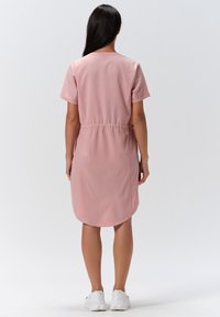 Rosa kurzärmliges Kleid mit geraffter Taille, glatter Textur und abgerundetem Saum. Getragen mit weißen Sportschuhen.