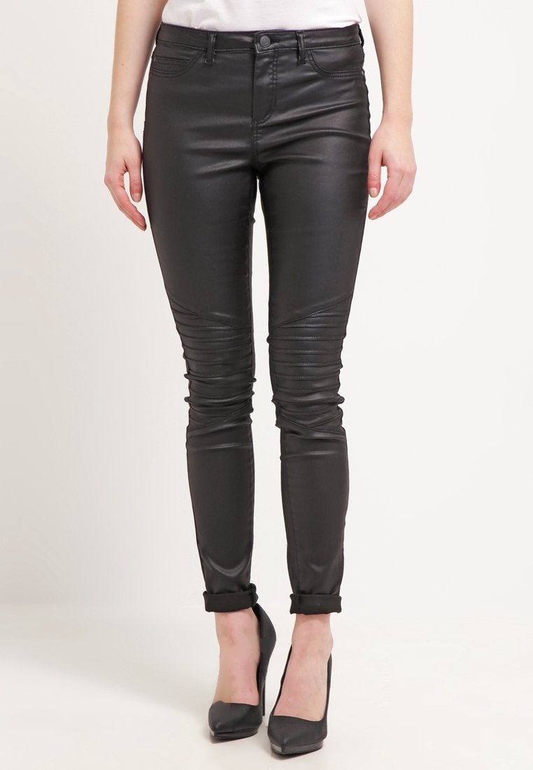Femme portant un pantalon en faux cuir noir ajusté avec des détails aux genoux et des chaussures à talons hauts noires sur un fond uni.